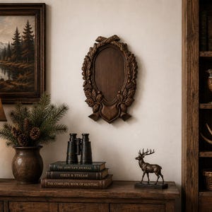 May include: A dark wooden wall plaque with an oval center, surrounded by a carved wreath design. A bronze deer statue sits on a wooden surface with books and binoculars. A framed landscape painting and a lamp are also visible.