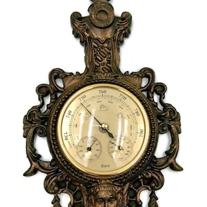 May include: An antique-style wall barometer in bronze tones, with a circular dial displaying weather symbols and pressure readings. The decorative frame includes scrollwork and a sculpted face. The dial is marked with the word "Baro".