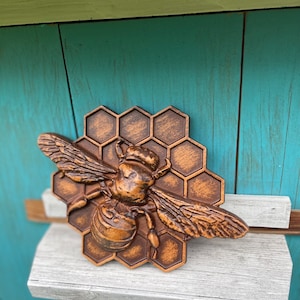 May include: A wooden bee sculpture with a honeycomb design, painted in a brown and gold color scheme. The bee is in a 3D style and has detailed wings and legs. The sculpture is mounted on a wooden shelf.