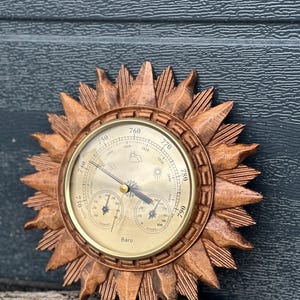 May include: A wooden barometer in a sunburst design. The barometer has a brass-coloured face with a needle and weather indicators. The outer frame is carved with sun rays. The barometer is placed on a wooden surface.