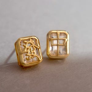 May include: A pair of gold-coloured square stud earrings. One earring has an etched design, the other a grid pattern. Both have a clear, faceted centre. The earrings are shown against a neutral background.