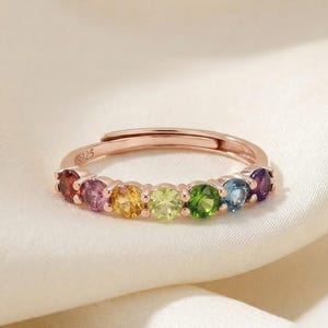 May include: Rose gold adjustable ring featuring a row of colorful gemstones in a rainbow spectrum. The gemstones include red, pink, yellow, green, blue, and purple. The ring is set on a white satin background.