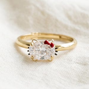 May include: A gold-toned ring featuring a central oval-shaped clear gemstone shaped like a cat face. The design includes a small red enamel bow detail on the upper right side and thin black enamel lines representing whiskers on the sides of the stone.