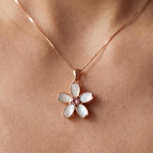 May include: A delicate necklace featuring a flower pendant. The pendant has a gold-colored frame with white petals and a pink center stone. The chain is a fine, gold-colored link, creating an elegant and feminine accessory.