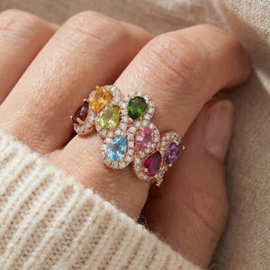 May include: A rose gold ring featuring a cluster of colorful gemstones, including yellow, green, blue, pink, and purple, each surrounded by a halo of small, clear stones. The ring is worn on a finger, with a cream-colored sweater in the background.