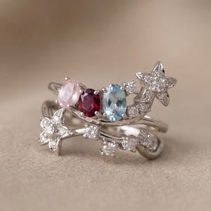 May include: A silver ring with three oval gemstones in pink, red, and blue, accented with star and floral designs. The ring has multiple bands and is set with small, clear stones.