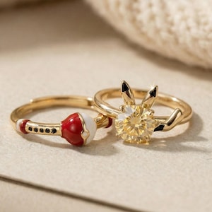 May include: Two gold rings. One ring features a red and white design with black accents. The other ring has a yellow gemstone and a bunny ear design. Both rings are on a neutral surface.