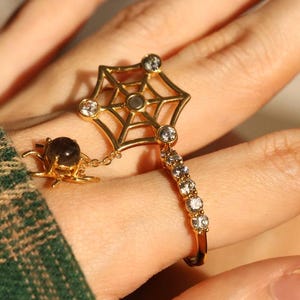May include: A gold-colored ring featuring a spiderweb design with clear gemstones and a small spider charm. The ring has a delicate chain connecting the spider charm to the web design. The ring is worn on a finger.