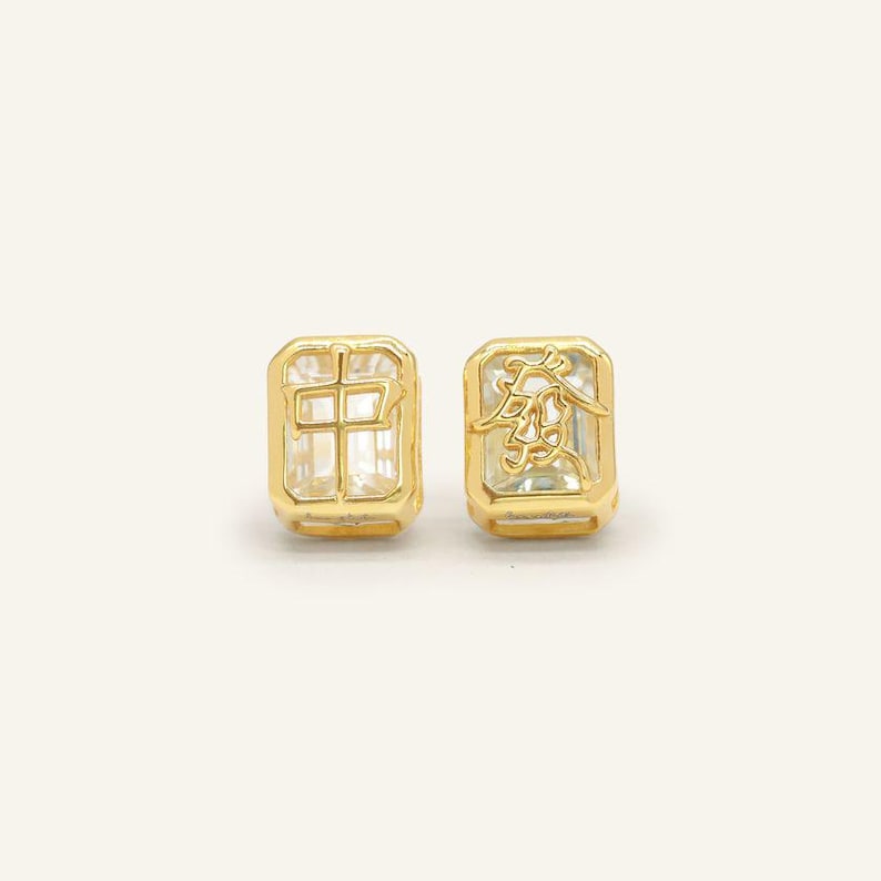 Peut inclure: Deux boucles d'oreilles dor&eacute;es avec des centres rectangulaires et transparents en forme de gemmes. L'une porte un caract&egrave;re chinois, l'autre un motif orn&eacute;. Fond clair et uni.