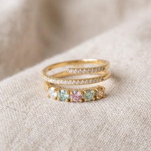 May include: A gold bypass ring featuring two bands, one encrusted with small clear stones and the other set with five round-cut gemstones in white, green, pink, green, and yellow, creating a multi-colored arrangement on a polished gold metal base.