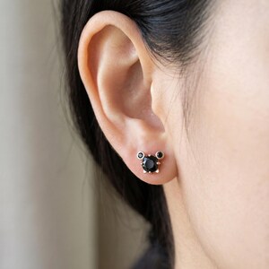 May include: A person's ear with a pair of black stud earrings. The earrings have a gold-coloured setting with a large, round black stone in the centre and two smaller black stones on either side.