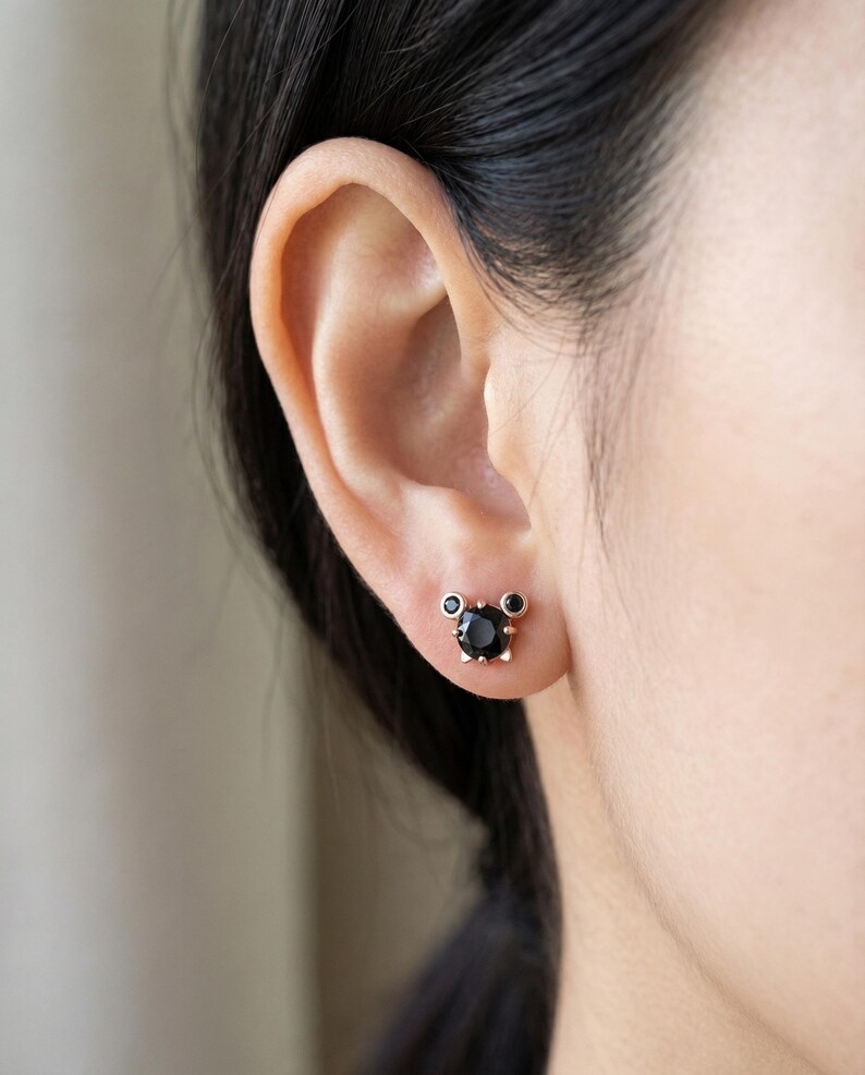 May include: A person's ear with a pair of black stud earrings. The earrings have a gold-coloured setting with a large, round black stone in the centre and two smaller black stones on either side.