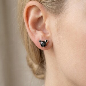 May include: A close-up of an ear with a black gemstone stud earring. The earring features a large, round black stone, with two smaller black stones above. The setting is a light metal colour.