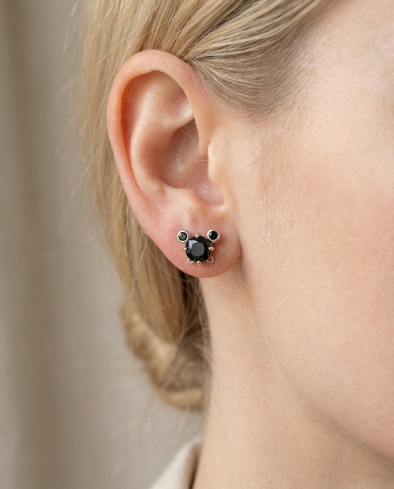 May include: A close-up of an ear with a black gemstone stud earring. The earring features a large, round black stone, with two smaller black stones above. The setting is a light metal colour.