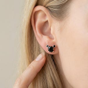 May include: A pair of black gemstone stud earrings. The earrings are set in a gold-coloured metal and feature a large, round black stone in the centre, with two smaller black stones on either side. The earrings are on a person's ear.