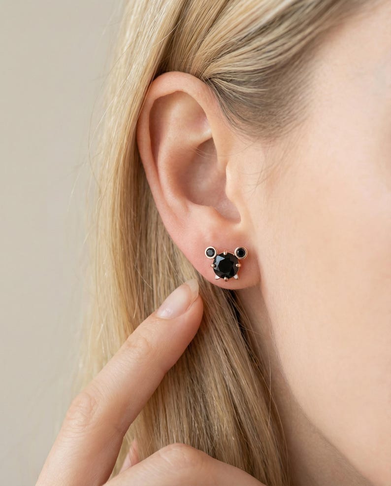 May include: A pair of black gemstone stud earrings. The earrings are set in a gold-coloured metal and feature a large, round black stone in the centre, with two smaller black stones on either side. The earrings are on a person's ear.