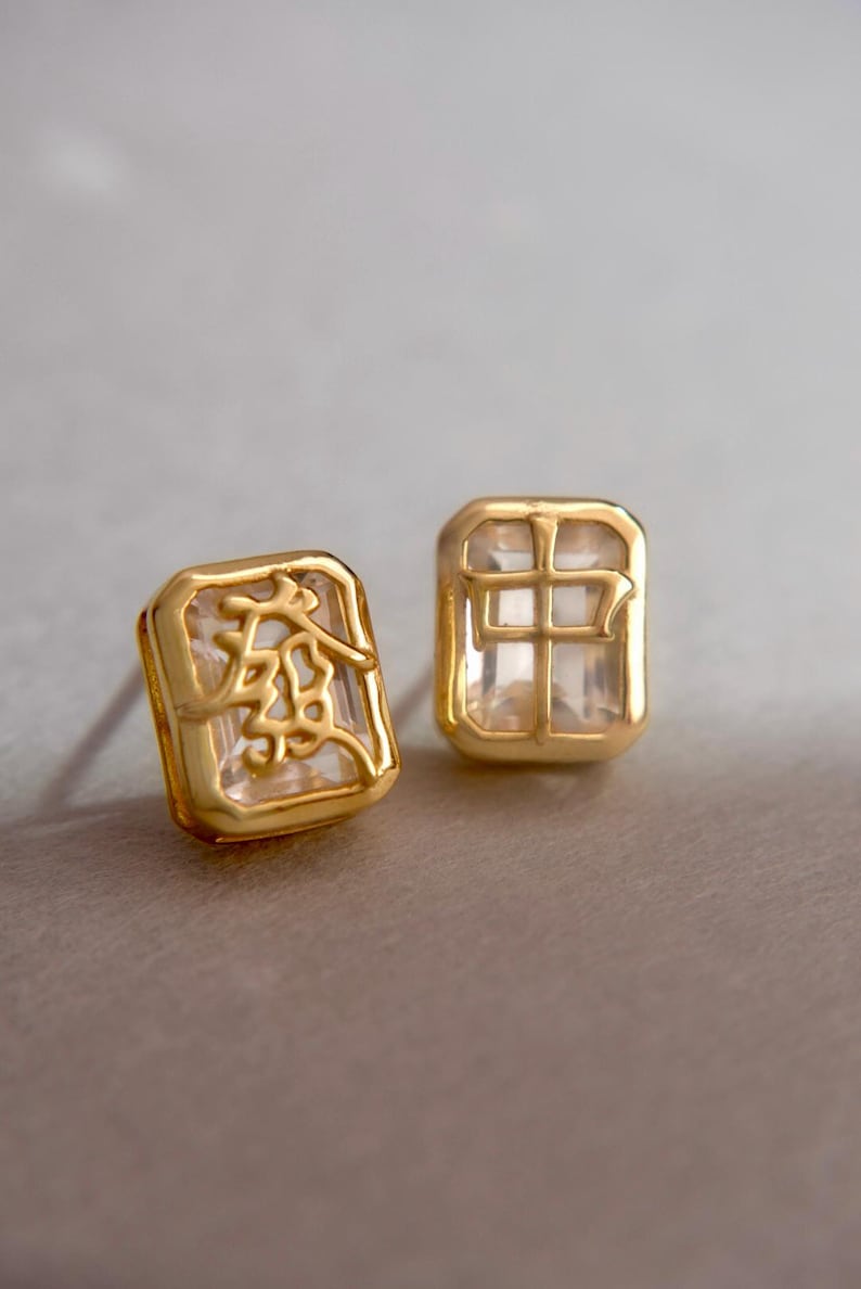 May include: A pair of gold-coloured square stud earrings. One earring has an etched design, the other a grid pattern. Both have a clear, faceted centre. The earrings are shown against a neutral background.