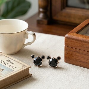 May include: Silver-tone stud earrings with a black gem centre and two smaller black gems above, styled like a cartoon character. The earrings are on a white surface next to a book titled "Disney Fine Art".