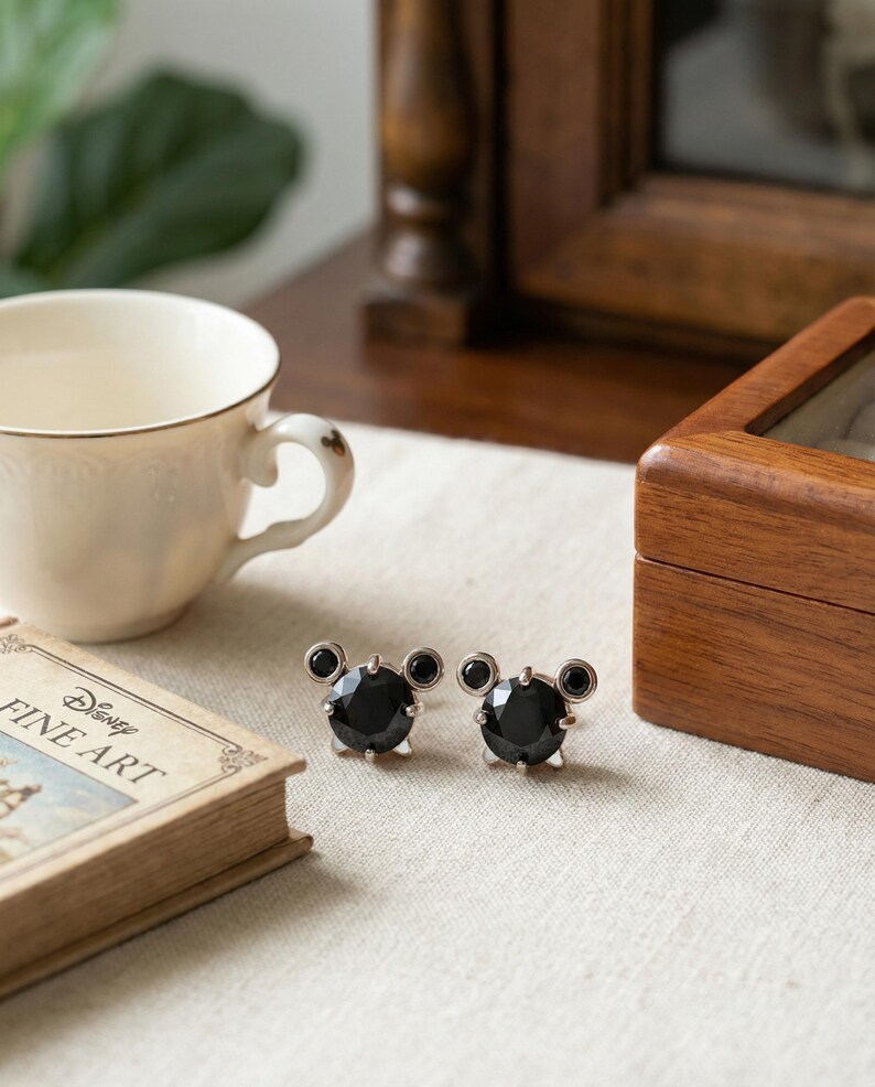May include: Silver-tone stud earrings with a black gem centre and two smaller black gems above, styled like a cartoon character. The earrings are on a white surface next to a book titled "Disney Fine Art".