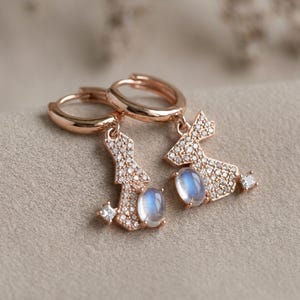 Può includere: Orecchini a cerchio in oro rosa con un design pendente. Ogni orecchino presenta una piccola forma geometrica scintillante incastonata con minuscole pietre trasparenti e una pietra ovale bluastra. Gli orecchini sono esposti su una superficie dai toni neutri.