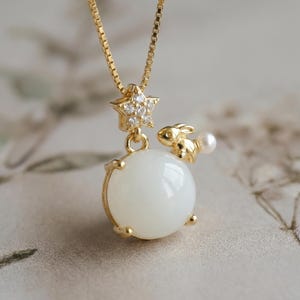 Puede incluir: Un collar dorado con un colgante de orbe blanco, un pequeño conejo dorado y una perla. El collar también tiene un dije en forma de estrella con pequeñas piedras transparentes. La cadena es una delicada cadena de caja.