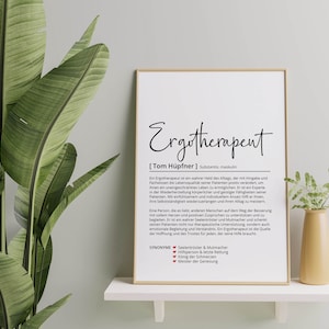 May include: A framed print with a white background and black text. The text reads "Ergotherapent [Tom H&uuml;pfner] Substantiv, maskulin" and a definition of the word. The print is on a white shelf with a green plant and a small vase with a flower on either side.