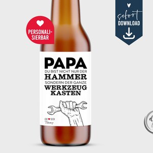 Peut inclure: Une bouteille de bière brune avec une étiquette blanche portant le mot "PAPA" en gras noir. L'étiquette contient également le texte "DU BIST NICHT NUR DER HAMMER SONDERN DER GANZE WERKZEUG KASTEN" et un dessin d'une main tenant une clé. Un autocollant rouge en forme de cœur est à gauche.