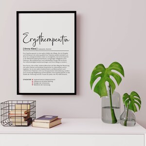 May include: A black and white framed print with the word "Ergotherapeutin" in bold, followed by the definition of the word in German. The definition is in a smaller font size and includes the name "Anna Klee". The print is hanging on a white wall above a white dresser with a black wire basket and books on top.