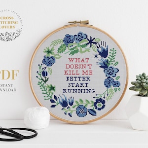May include: A cross stitch pattern featuring a floral wreath with blue flowers and green leaves. The text inside the wreath reads "What doesn't kill me better start running".