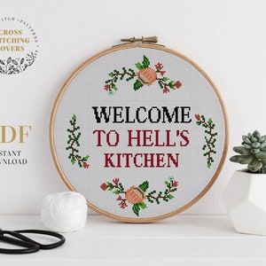 Pode incluir: Um aro de bordado branco com um desenho de ponto cruz que diz "Bem-vindo à cozinha do inferno" com detalhes florais. O aro está sobre uma superfície branca com uma pequena planta suculenta em vaso.