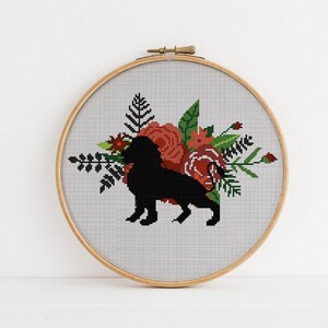 Puede incluir: Una silueta negra de un perro con un arreglo floral de flores rojas y verdes en un aro de bordado de madera.