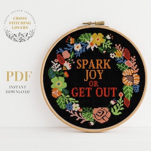 以下が含まれることがあります： 黒い刺繍枠に、花輪のデザインと「Spark Joy or Get Out」というテキストが書かれています。