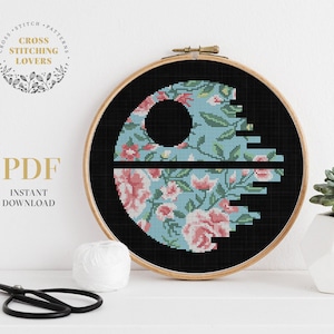 May include: A cross-stitch pattern in a wooden hoop features a floral design within a black circle, resembling a Death Star. The floral pattern includes pink roses and green leaves on a light blue background. The text "PDF INSTANT DOWNLOAD" is visible.