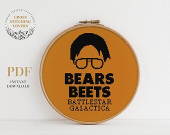 May include: A round, orange-coloured embroidery hoop with a black and white cross-stitch design. The design features a cartoon character with glasses and the text "Bears Beets Battlestar Galactica".