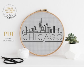 以下が含まれることがあります： シカゴのスカイラインの白黒のクロスステッチパターンで、下に「CHICAGO」という文字があります。パターンは木製の刺繍枠に表示されています。
