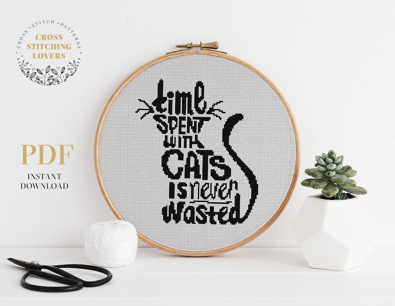 May include: A white embroidery hoop with a black and white cross stitch design that reads "Time spent with cats is never wasted".