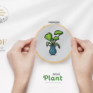 May include: A close-up of a cross stitch pattern of a small green plant in a blue vase. The pattern is in a wooden embroidery hoop and is being held by a person's hands. The text "mini Plant" is below the hoop.