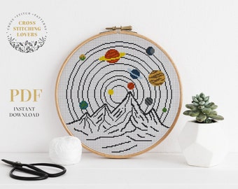 May include: A cross stitch pattern featuring a stylized depiction of a mountain range with planets orbiting a sun in the background. The pattern is displayed in a wooden embroidery hoop.