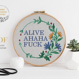 Puede incluir: Un patrón de punto de cruz con una corona de flores con el texto "ALIVE AHAHA FUCK" en letras azules. El patrón se muestra en un aro de bordado de madera.