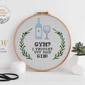 Könnte beinhalten: Ein weißer Stickrahmen mit einem Kreuzstich-Motiv einer Gin-Flasche und einem Glas. Das Motiv ist von einem Kranz aus grünen Blättern und rosa Blüten umgeben. Der Text "GYM? I THOUGHT YOU SAID GIN!" ist in schwarzem Garn gestickt.