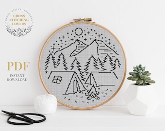 May include: A black and white cross stitch pattern of a mountain scene with a tent, campfire, and stars. The pattern is in a wooden embroidery hoop on a white surface.
