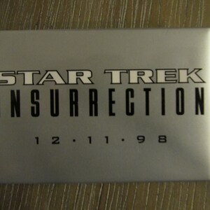 May include: A rectangular, silver-colored Star Trek: Insurrection collectible with black text. The title is in a bold, stylized font, with the date "12.11.98" below. The item is likely a promotional item or memorabilia.