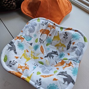 May include: A set of two orange and white fabric pot holders with a woodland animal print. The pot holders are quilted and have a square and round shape.
