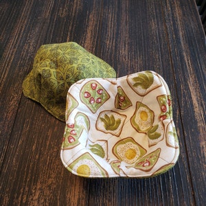 May include: Two fabric bowls, one green with a geometric pattern and one white with a pattern of avocado toast and strawberries. The bowls are on a dark wood surface.
