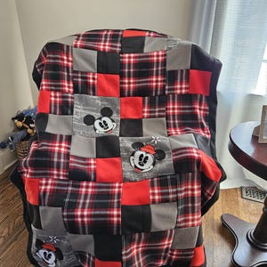May include: A red, black, and gray plaid fleece blanket with embroidered Mickey Mouse and Minnie Mouse faces. The blanket is folded and sitting on a chair.