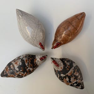 May include: Four ceramic bird sculptures arranged in a circle. The birds are teardrop-shaped and feature a range of colours: white with crackle lines, brown, and black with brown and white patterns. Each bird has a red beak.