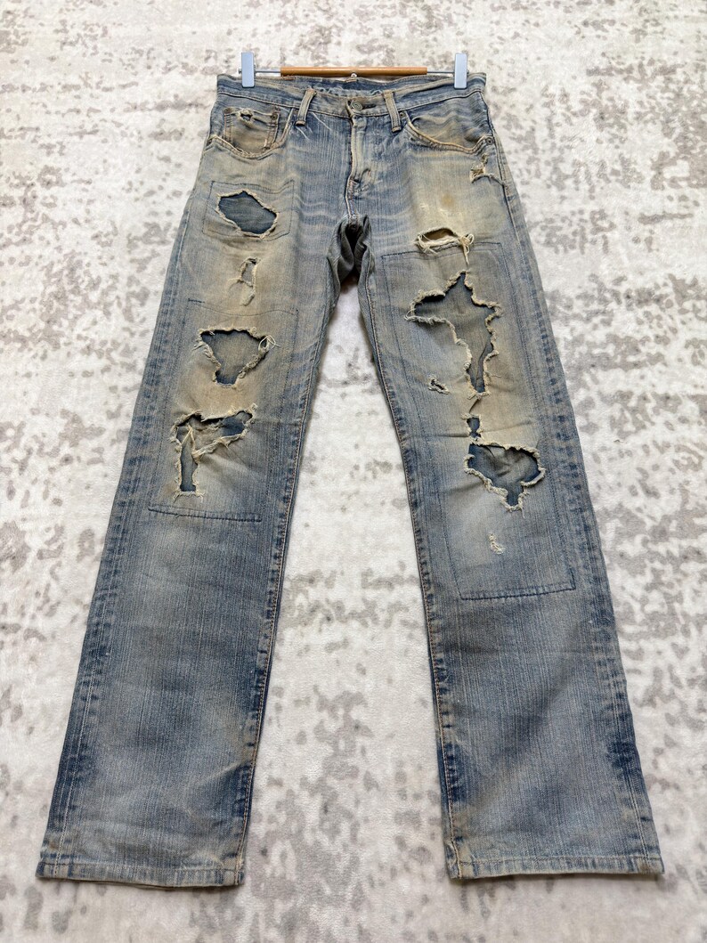 Puede incluir: Vaqueros de mezclilla azul claro con detalles desgastados, con grandes rasgaduras y parches. Los vaqueros cuelgan de una percha de madera, mostrando una est&eacute;tica vintage y desgastada. Los vaqueros tienen un dise&ntilde;o cl&aacute;sico de cinco bolsillos.