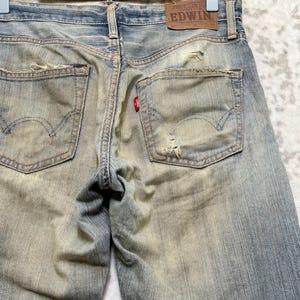 Puede incluir: Vaqueros de mezclilla azul descolorido con detalles desgastados, incluyendo rasgaduras y parches desgastados. Los bolsillos traseros presentan un dise&ntilde;o cl&aacute;sico, y una peque&ntilde;a etiqueta roja es visible. Un parche de cuero marr&oacute;n con la marca "EDWIN" est&aacute; cosido en la cintura.