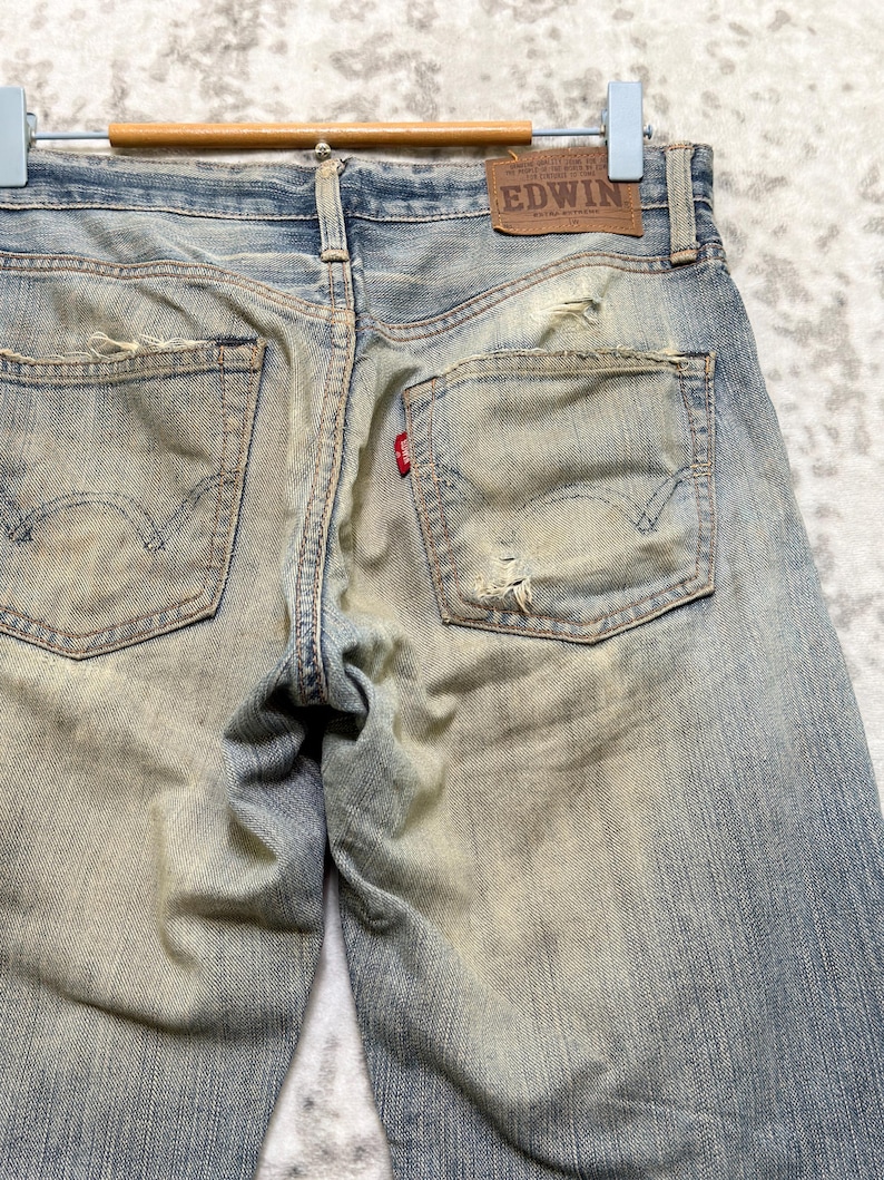 Puede incluir: Vaqueros de mezclilla azul descolorido con detalles desgastados, incluyendo rasgaduras y parches desgastados. Los bolsillos traseros presentan un dise&ntilde;o cl&aacute;sico, y una peque&ntilde;a etiqueta roja es visible. Un parche de cuero marr&oacute;n con la marca "EDWIN" est&aacute; cosido en la cintura.
