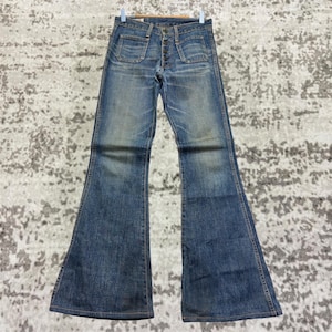 Puede incluir: Vaqueros vintage de mezclilla azul con campana. Los vaqueros tienen bragueta de botones, dos bolsillos delanteros y dos traseros. La mezclilla está descolorida y desgastada, lo que le da a los vaqueros un aspecto retro clásico. Los vaqueros están colgados de una percha de madera.